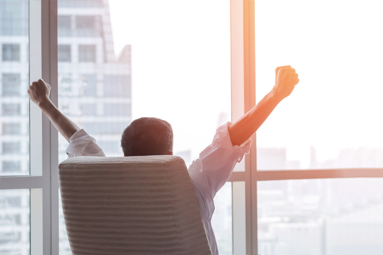 happy businessman in office raising fists looking forward to city building urban scene through glass window