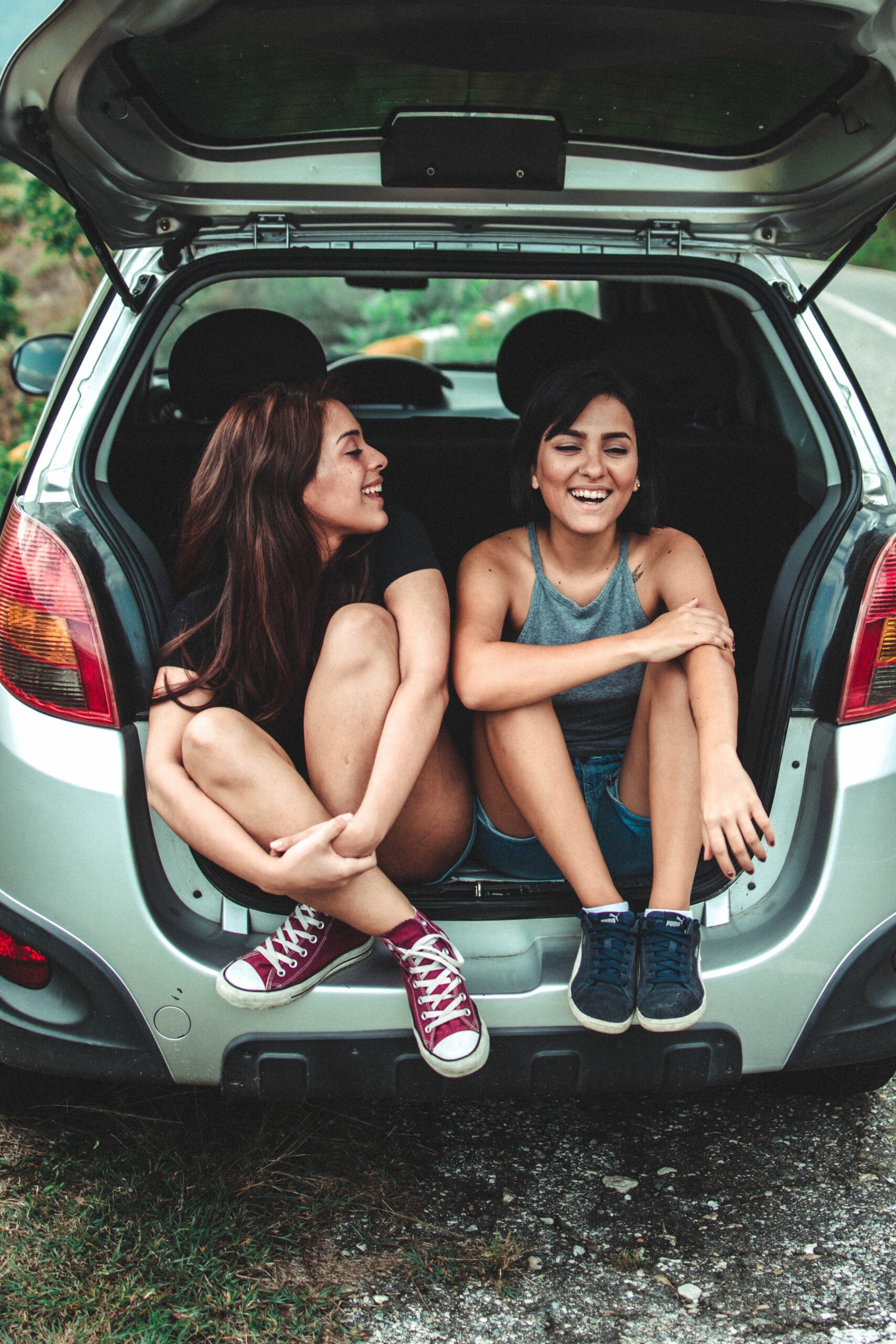 two girls sitting in the back of a car with the boot up and laughing with one another